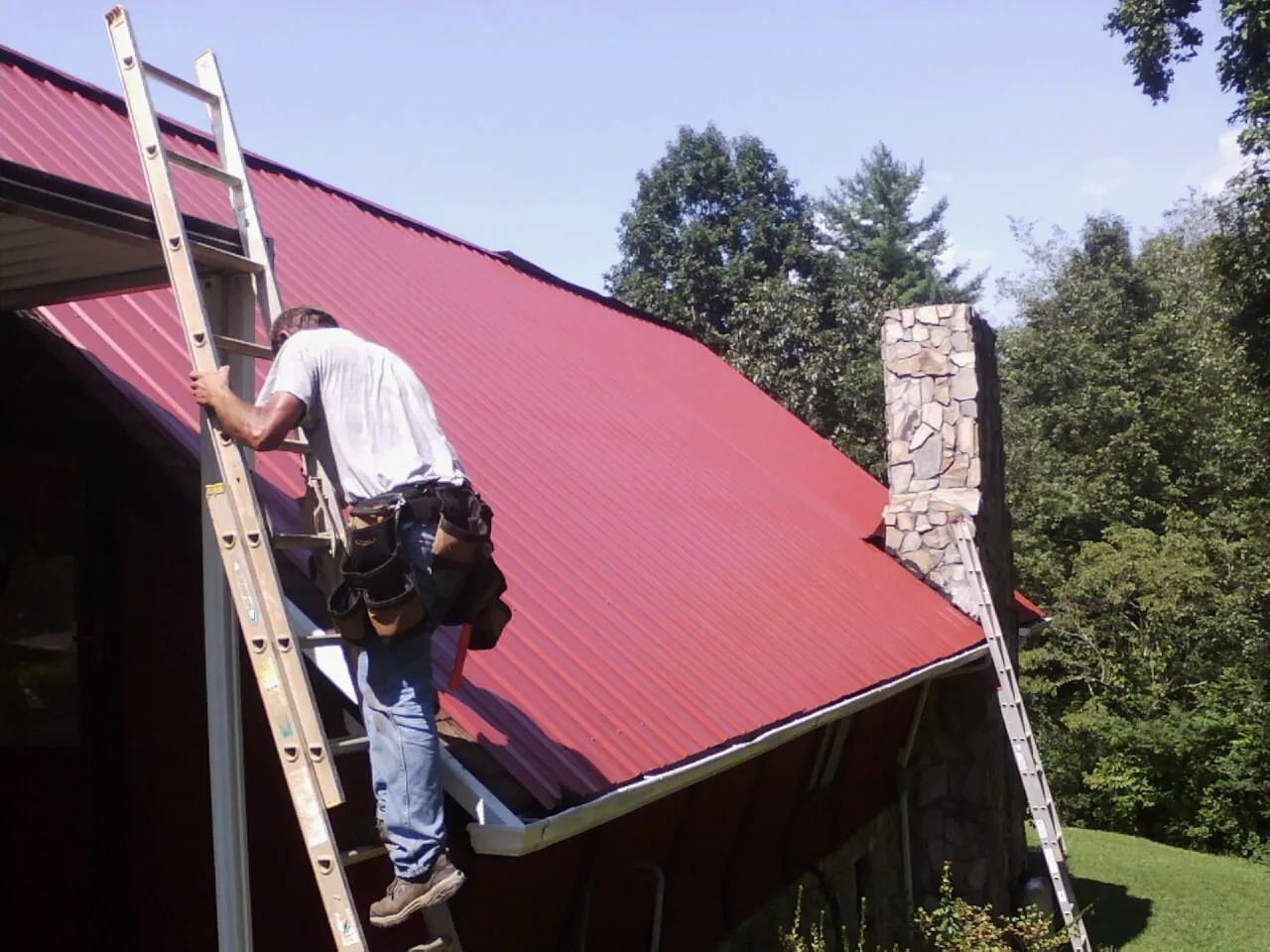 Roofer on a ladder beside a red metal roof and stone chimney — Soffit & Fascia Repair by Roofing Brothers in Germanton